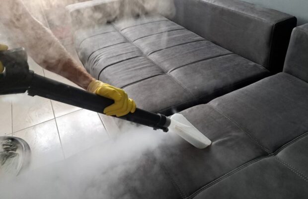 Curățarea unei canapele gri cu un aparat profesional cu abur, folosind o duză aplicată pe tapițerie.